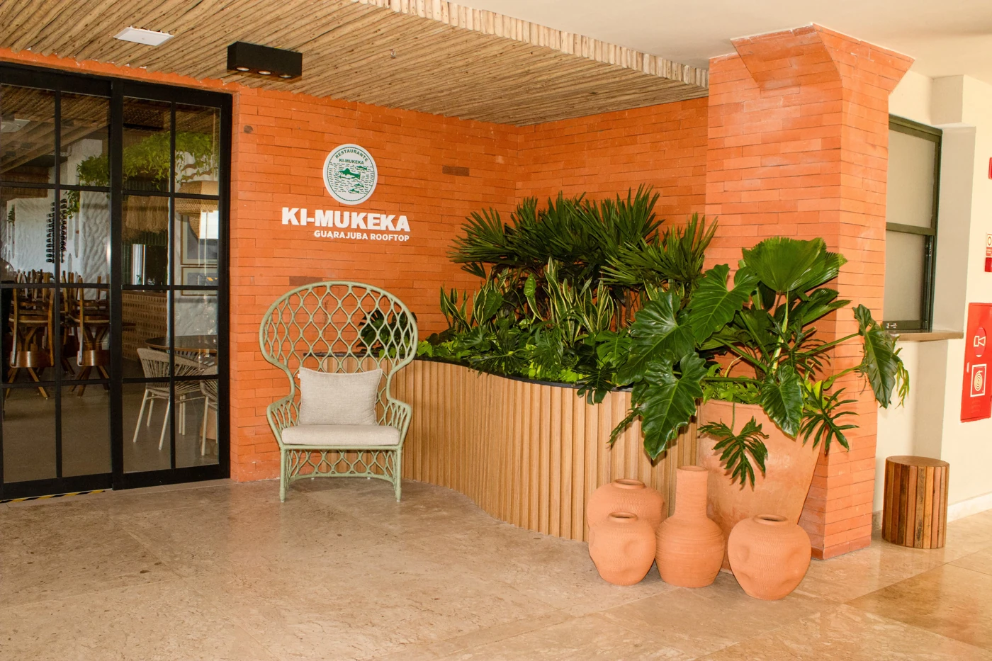 Entrada do Ki-Mukeka Guarajuba Rooftop, com parede de tijolos, plantas e mobiliário acolhedor